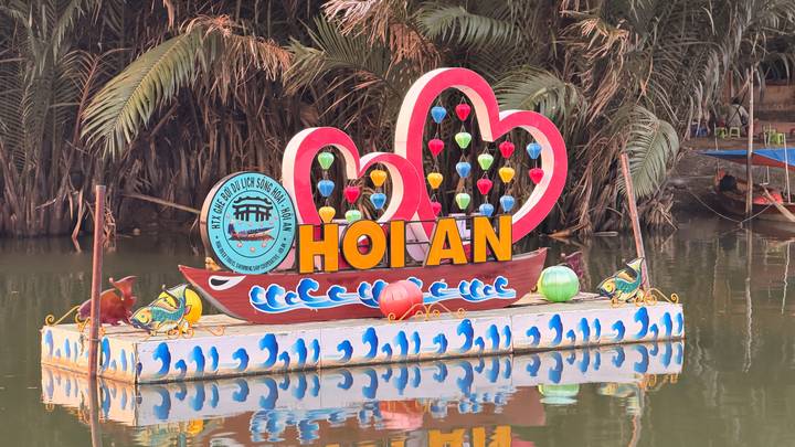 Colorful floating sign with lanterns spelling out “Hoi An” surrounded by palm foliage on a still canal.