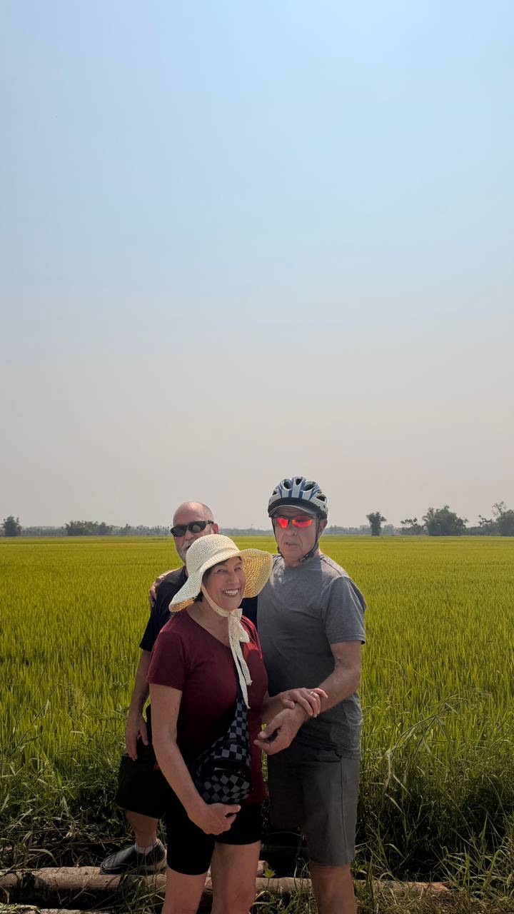 Three cyclists posing happily in front of a vast golden rice field under a hazy sky.