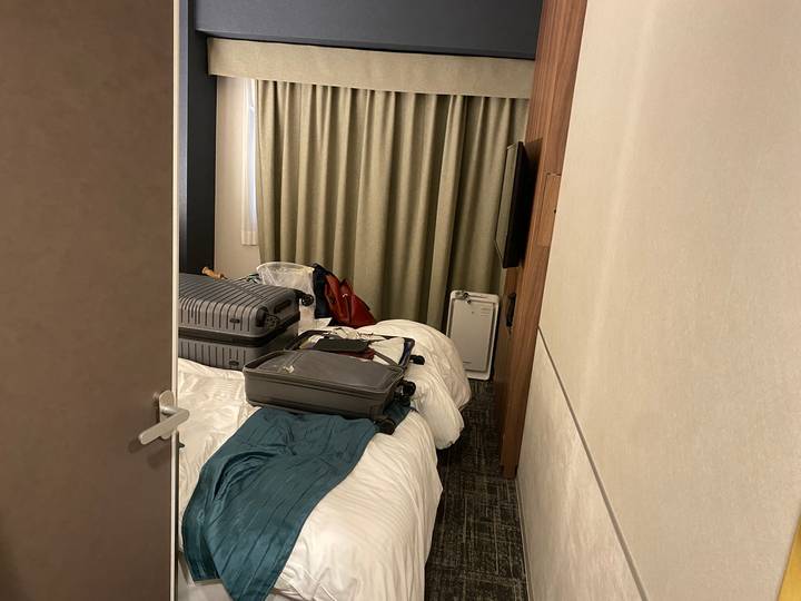 Small hotel room cluttered with open suitcases, bags and clothing on unmade beds.