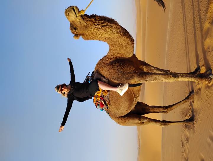 Happy traveler riding a camel across golden Sahara sand dunes under a clear blue sky.