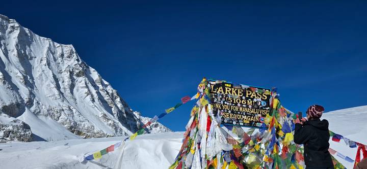 Colorful prayer flags flutter around the Larke Pass sign amid snowy Himalayan peaks while a trekker snaps a photo.