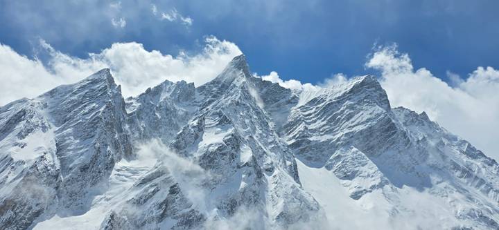 Jagged snow-covered Himalayan peaks pierce scattered clouds against a bright sky.