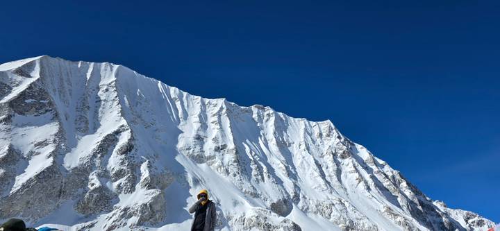Trekker bundled in winter gear stands below a towering icy Himalayan ridge.