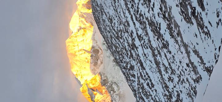 Golden sunlight ignites a snow-clad mountain summit above a rocky, snowy slope.