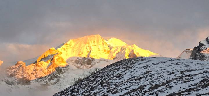 Panoramic sunset light bathes a dramatic snow-covered Himalayan peak above a ridgeline.