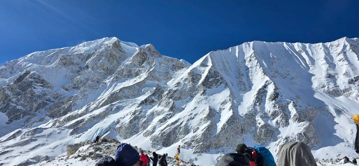Trekking group pauses beneath towering snowy cliffs and deep blue sky.