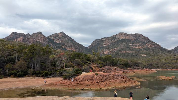 Visitors exploring rocky red granite headland with hazy mountain peaks behind calm bay.