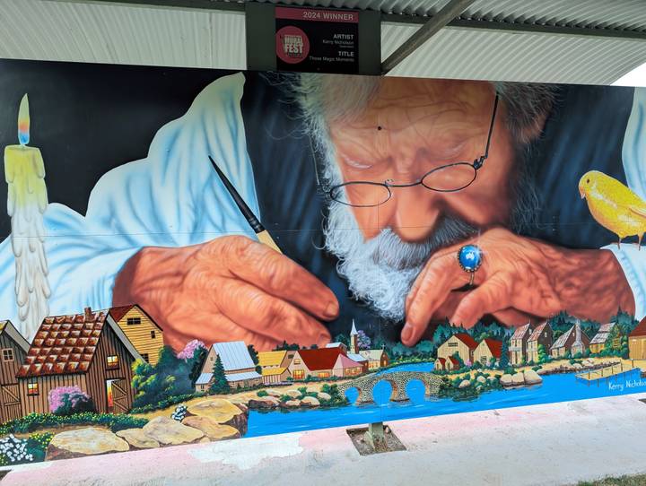 Colorful mural of an elderly man painting miniature houses, executed on a large exterior wall.