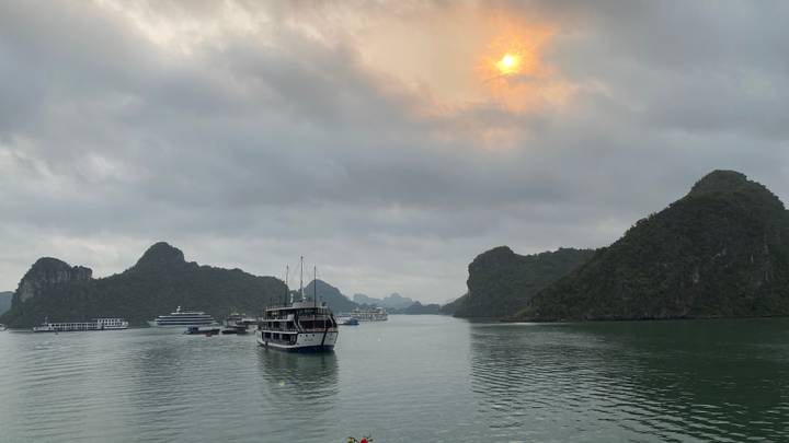 Cruise boats float among limestone karsts on a calm bay while the sun glows through a cloudy sky.