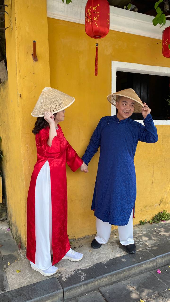 Two travelers in bright traditional Ao Dai and conical hats pose playfully against a vibrant yellow wall.