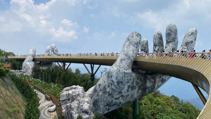 Crowds stroll across the famous Golden Bridge held by giant stone hands high above a forested valley.