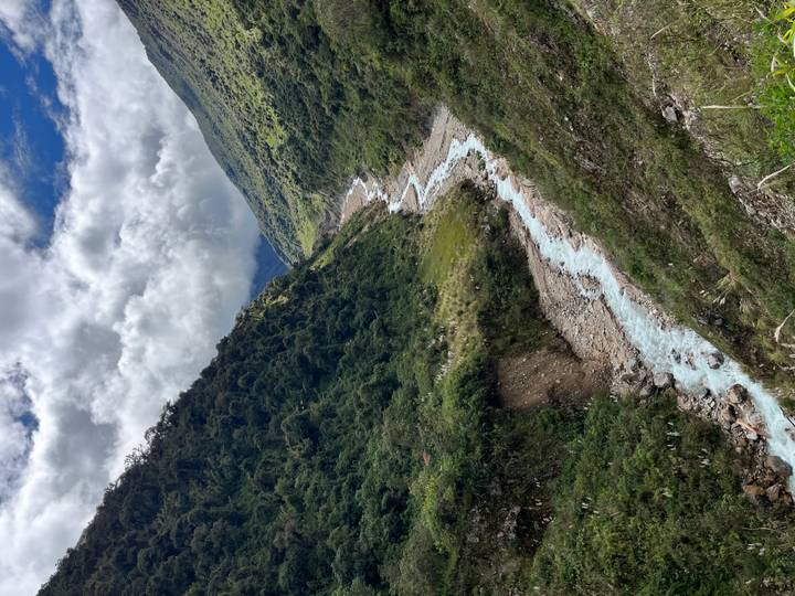 Pale blue glacial river winding through lush Andean valley under cloud-filled sky.
