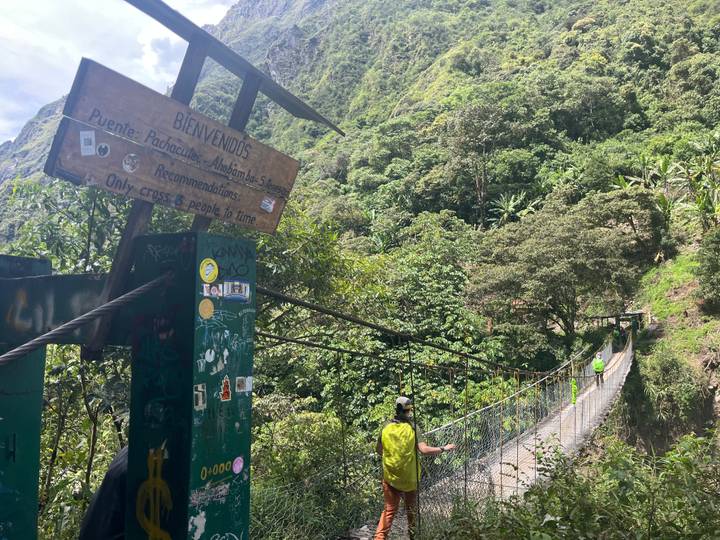 Hiker crossing narrow suspension bridge over jungle gorge with welcome signpost.
