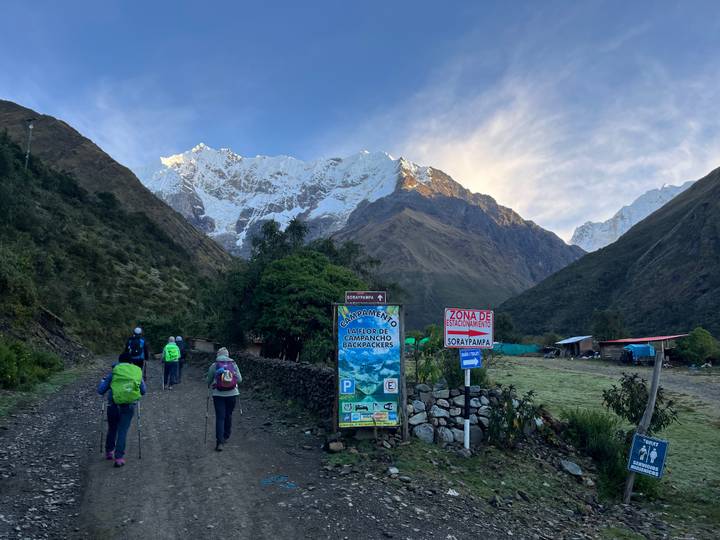 Trekkers with backpacks walk along a dirt road toward a dramatic snow-covered Andean peak under early morning light.