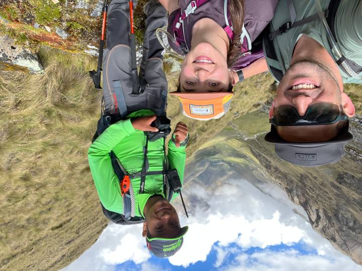 Three smiling hikers take a selfie on a high mountain pass with a lush green valley and cloudy peaks behind them.