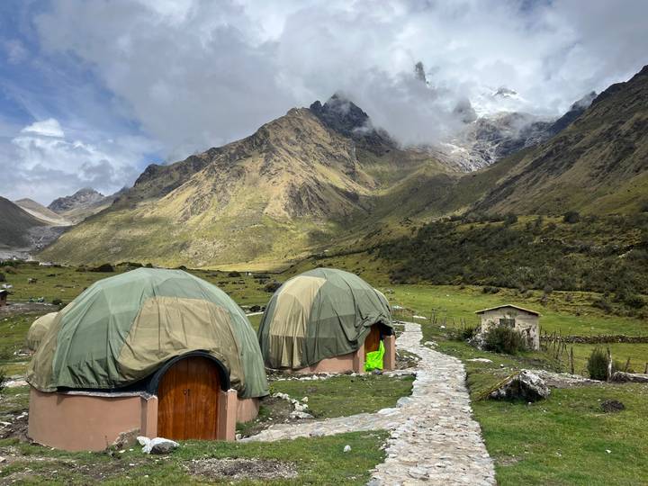 Round canvas-covered domes of a remote campsite sit on a grassy alpine meadow beneath rugged, cloud-shrouded peaks.