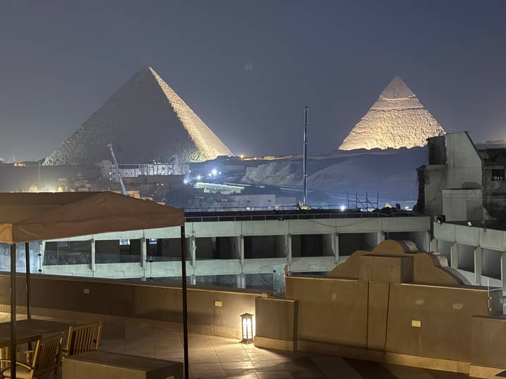 Nighttime view of the illuminated Great Pyramids of Giza rising behind urban rooftops.
