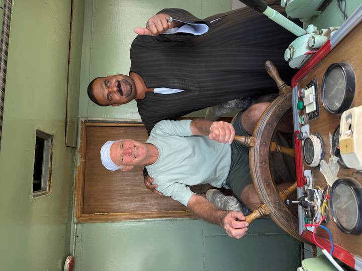 Two men, one steering a wooden ship's wheel, share a friendly moment aboard a riverboat.