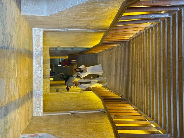 Tourists descend a long wooden staircase inside an ancient Egyptian tomb lined with hieroglyph-covered sandstone walls illuminated by yellow lights.