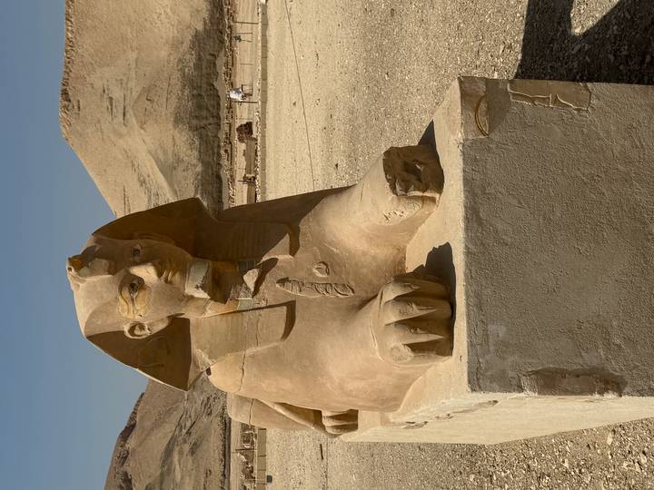 Sand-colored sphinx statue with human head and lion body positioned outdoors in a desert archaeological site.