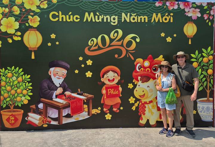 Smiling couple pose before a vibrant Tet New Year mural reading 'Chúc Mừng Năm Mới 2026'.