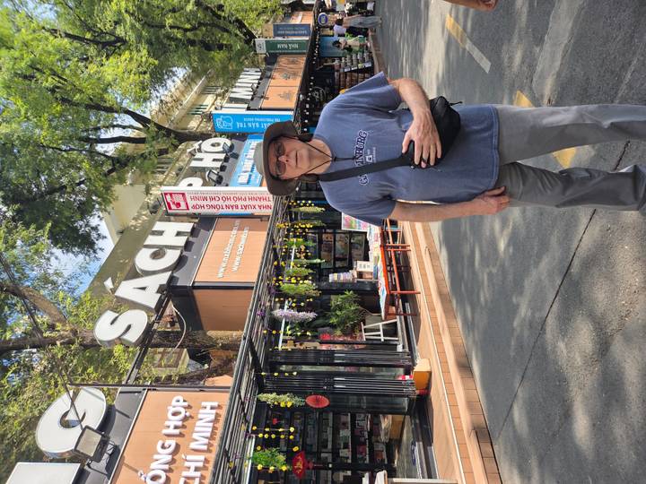 Tourist stands on a pedestrian book street lined with stalls under large 'Sách' signage and shady trees.