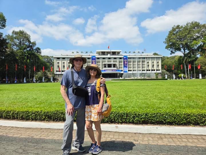 Couple stand on a lawn in front of Ho Chi Minh City’s Reunification Palace under a bright sky.