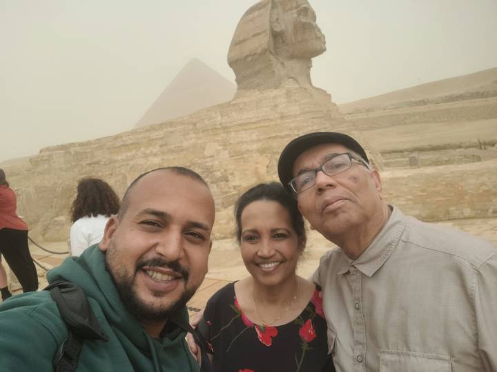 Selfie of three travellers smiling in front of the Sphinx and a pyramid on a dusty day.