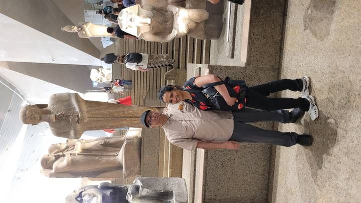 Couple standing among monumental ancient statues inside a museum with other visitors on steps.