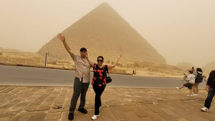 Couple celebrating with raised arms on the roadside in front of the Great Pyramid during a sandstorm.