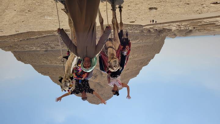 Two tourists on camels raising their hands with the Step Pyramid of Saqqara behind them and a camel handler in front.