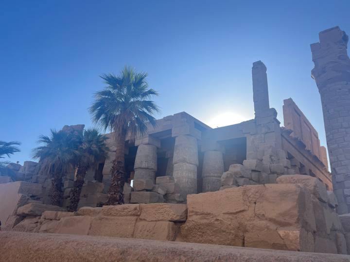 Ancient stone columns of Karnak Temple silhouetted by bright desert sun and palm trees.