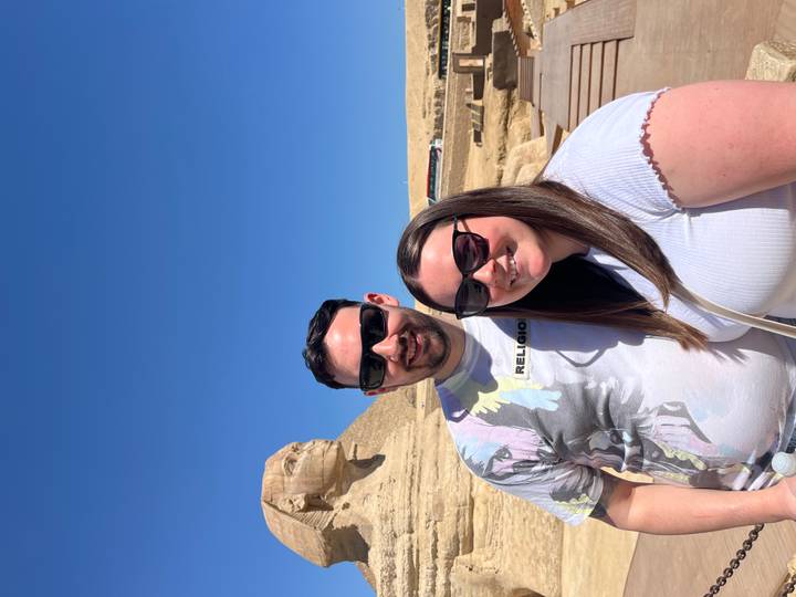 Couple wearing sunglasses posing beside the Sphinx and Pyramid under a clear blue sky.