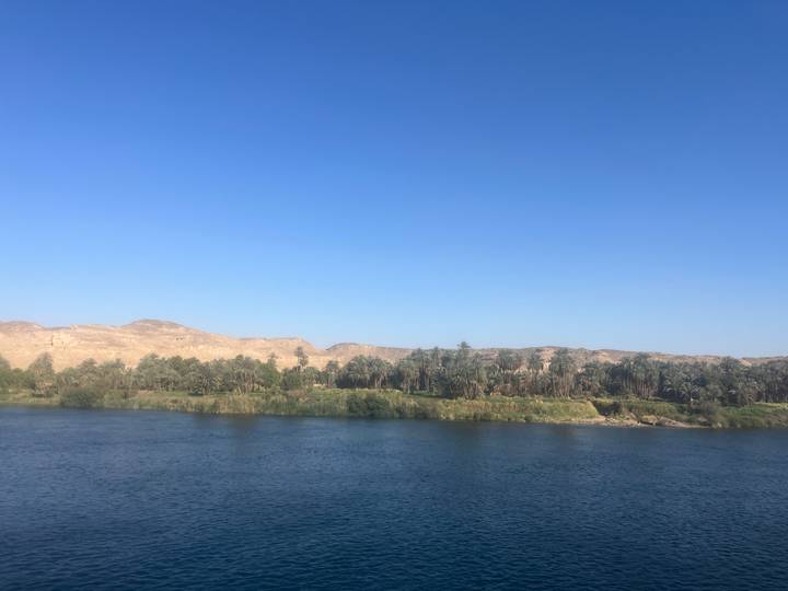 Blue waters of the Nile bordered by palm groves and desert hills under a cloudless sky.