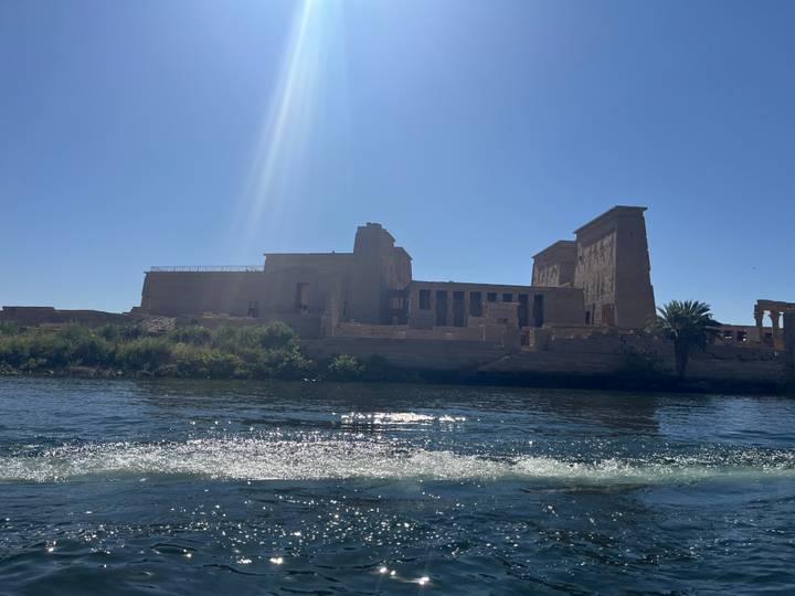 Ancient Philae Temple complex seen from a boat with bright sunlight creating a lens flare.