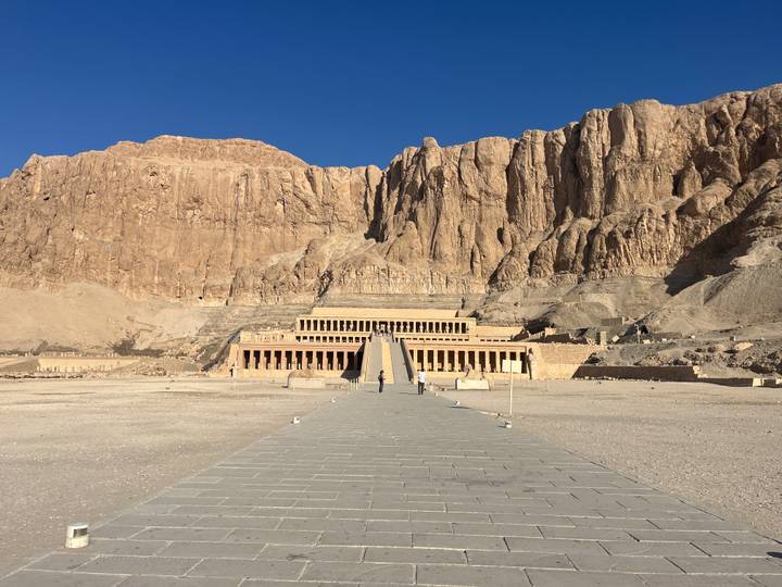 Wide stone pathway leading to Hatshepsut Temple backed by towering desert cliffs under clear blue sky.