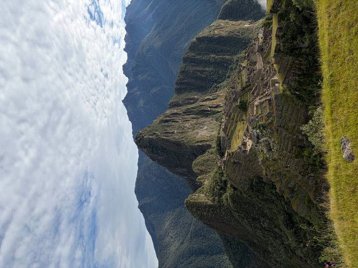 Vertical shot of Machu Picchu ruins under partly cloudy sky with forested peak towering behind.