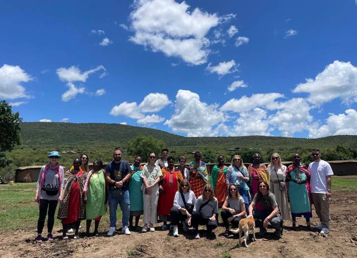 Travel group posing with Maasai women in front of green hills and traditional huts.