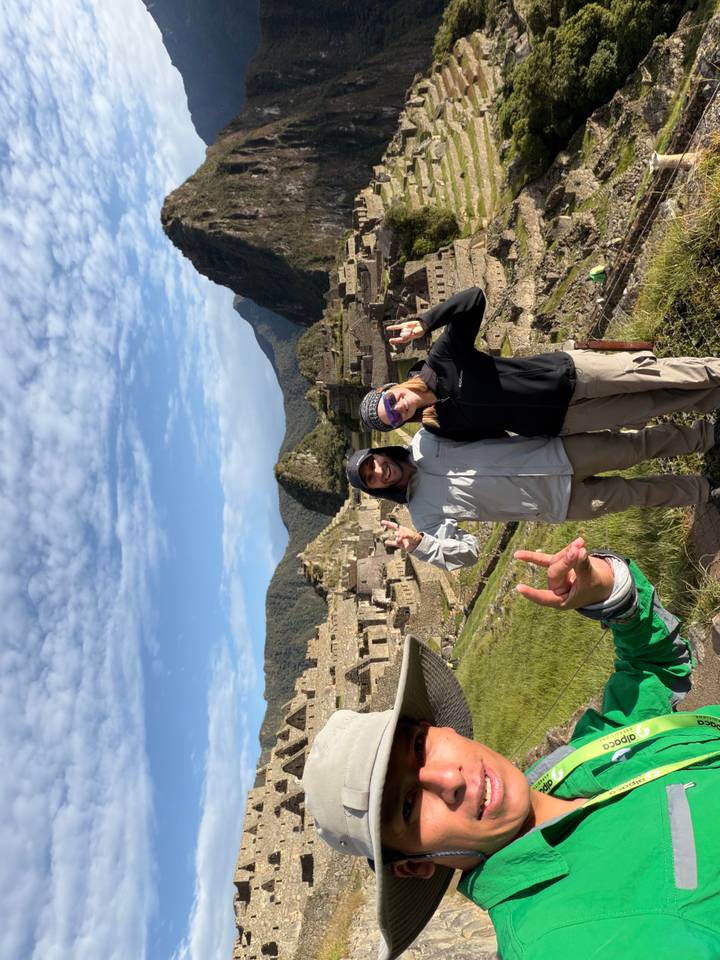 Two hikers posing with rock-on hand signs at Machu Picchu, another friend’s hand photobombing in foreground.