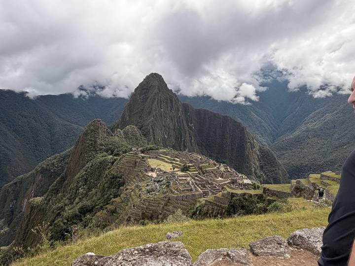 Iconic view of the Inca citadel of Machu Picchu surrounded by steep green mountains under a cloudy sky, with part of a visitor visible at the edge.