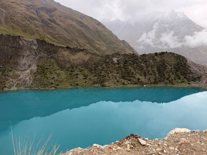 Turquoise glacial lake reflecting the surrounding rugged Andean slopes and misty clouds.