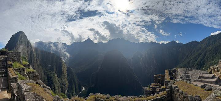 Wide panoramic shot of the lush green canyon and surrounding peaks beyond Machu Picchu ruins under dramatic skies.