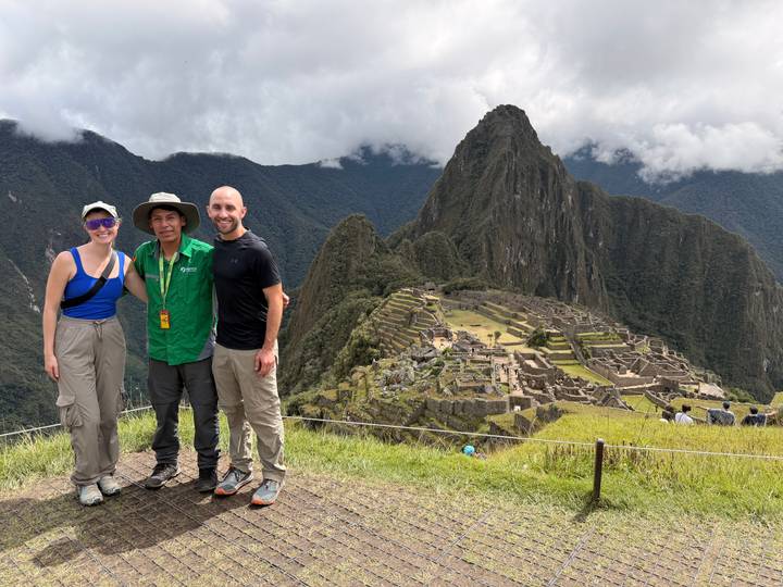 Three travellers pose smiling with Machu Picchu’s citadel and Huayna Picchu peak rising behind them.