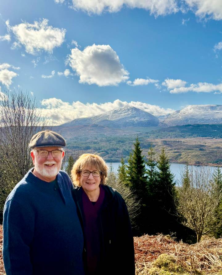 Smiling senior couple pose with snowy Scottish Highlands and a loch behind them on a crisp day.