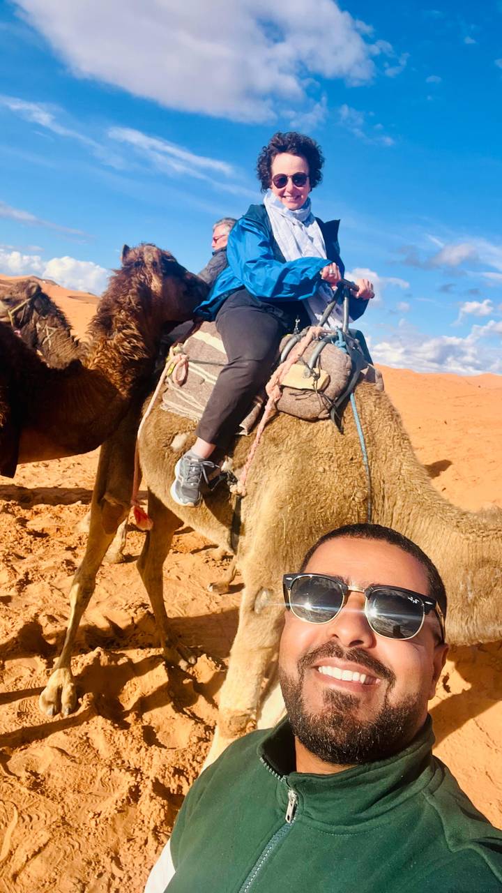 Close selfie partially showing a traveller riding a camel on sandy desert dunes.