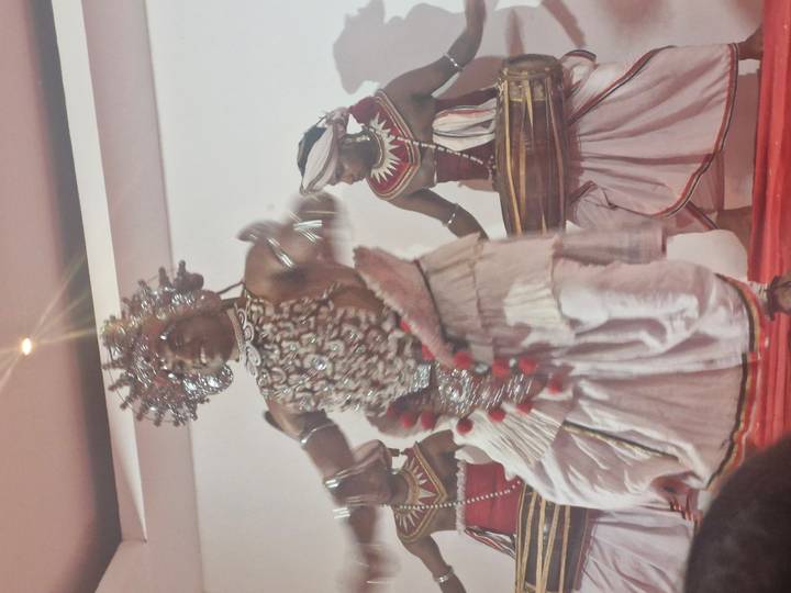 Traditional Kandyan dancers in elaborate costumes perform on an indoor stage.