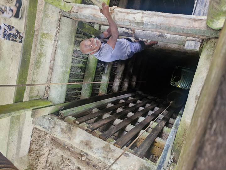A smiling man descends a wooden ladder into a deep traditional well.