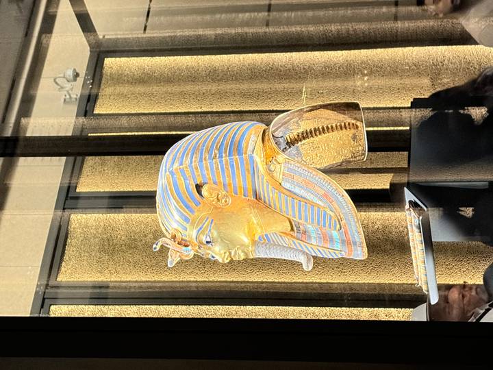 Golden mask of Tutankhamun displayed behind glass in a museum with visitors reflected