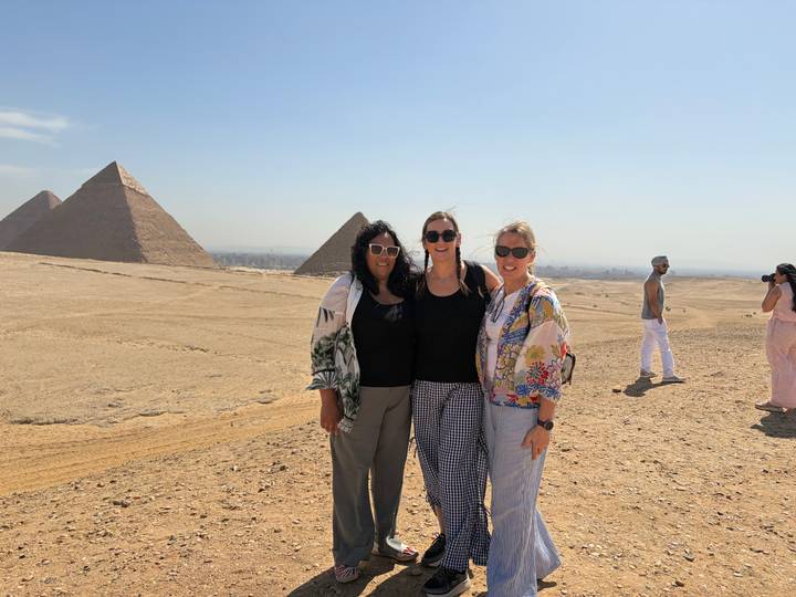 Three women pose smiling with pyramids rising from sandy desert plateau in bright sunlight