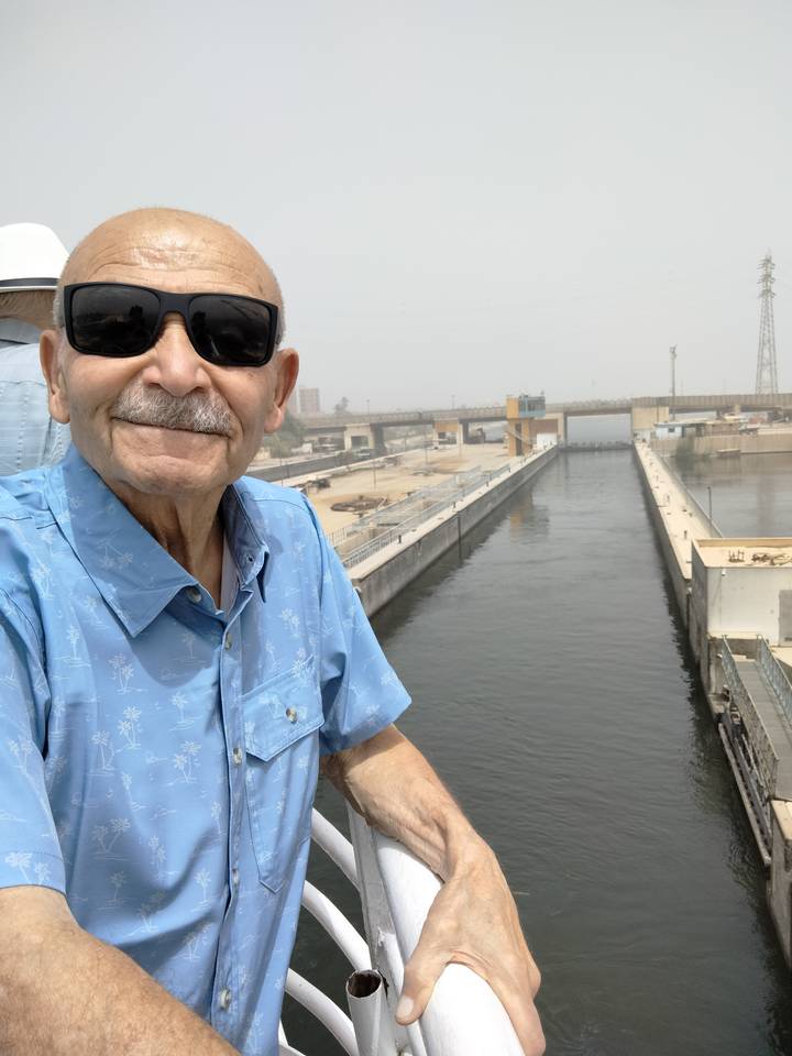 Smiling elderly man in sunglasses wearing a blue shirt taking a selfie on the deck of a boat beside a canal lock and calm water